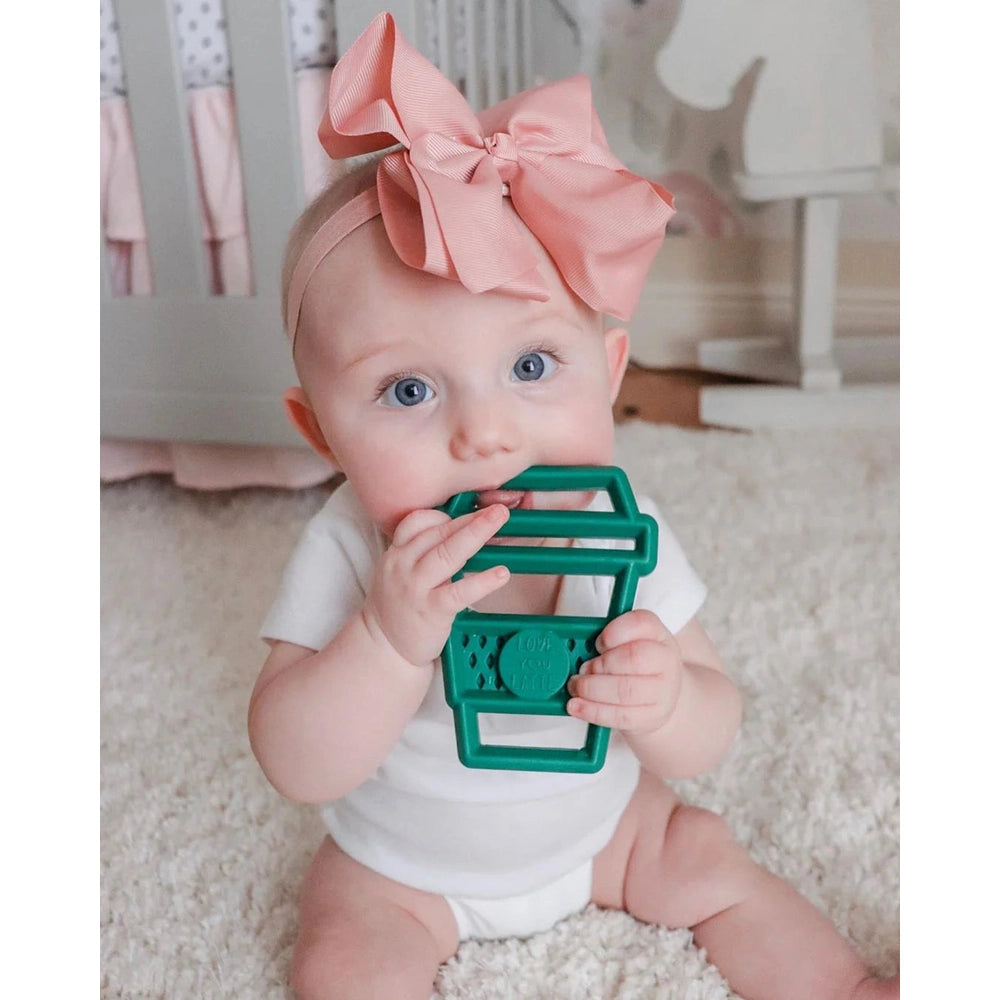 Itzy Ritzy silicone teether in teal-green held by baby with pink bow headband