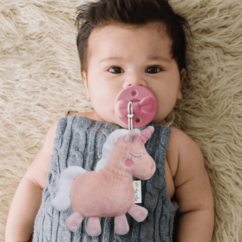 Itzy Ritzy Sweetie Pal pacifier with pink unicorn plush attached, baby lying on cream rug