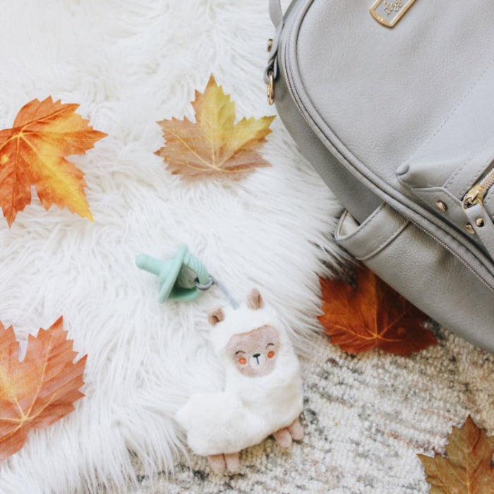 Itzy Ritzy Sweetie Pal pacifier with plush llama clip on white shag rug beside gray bag and fall leaves