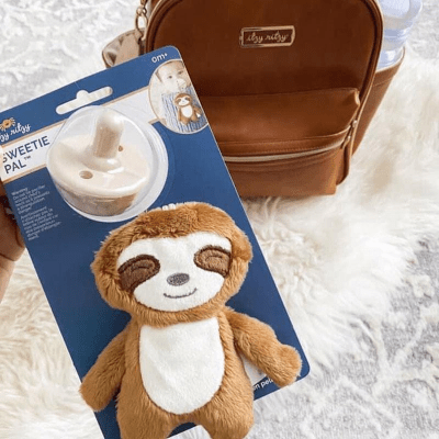 Itzy Ritzy Sweetie Pal pacifier with plush sloth on blue card; brown mini backpack in background.