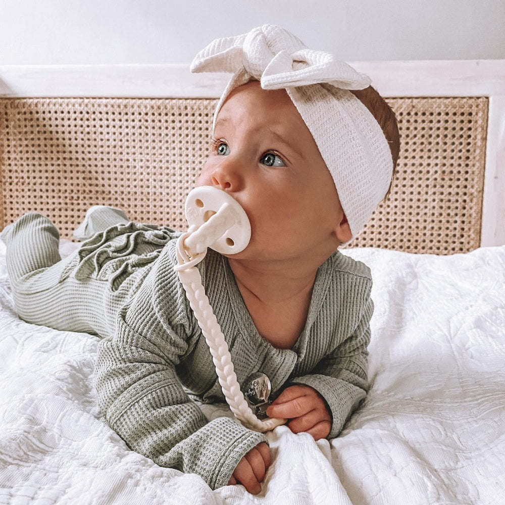Itzy Ritzy Sweetie Soother Pacifier 2-pack; baby on a white bed uses a white pacifier with braided teether.