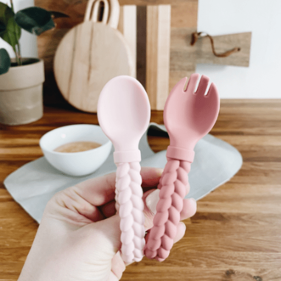 Itzy Ritzy Sweetie Spoon & Fork Set in pastel pink with braided handles, held in hand to show comfy grip.