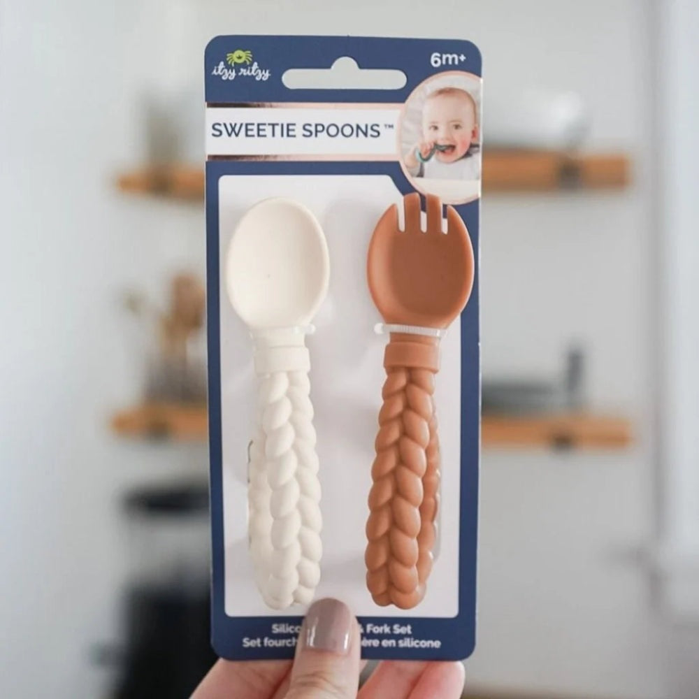 Itzy Ritzy Sweetie Spoon & Fork Set, white spoon and brown fork with braided silicone handles, held in hand.