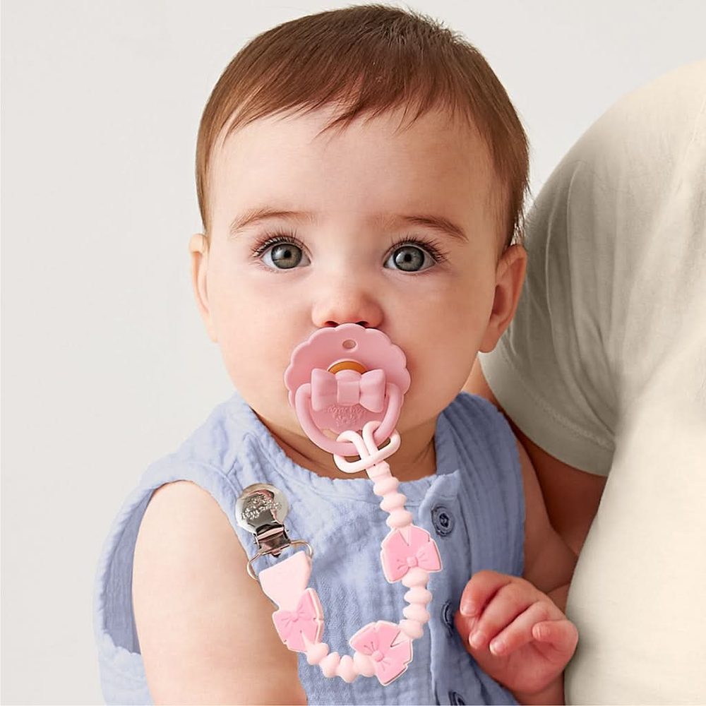 Itzy Ritzy Sweetie Strap Plus Bow pacifier clip attached to a pink pacifier on baby