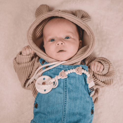 Itzy Ritzy Sweetie Strap Plus pacifier clip on a baby wearing denim overalls and a tan bear hoodie