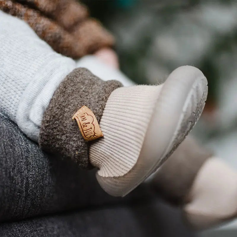 Jan & Jul Cozy Sock Shoes Oat, baby's foot snug in soft beige knit sock with cushioned sole and leather tag.