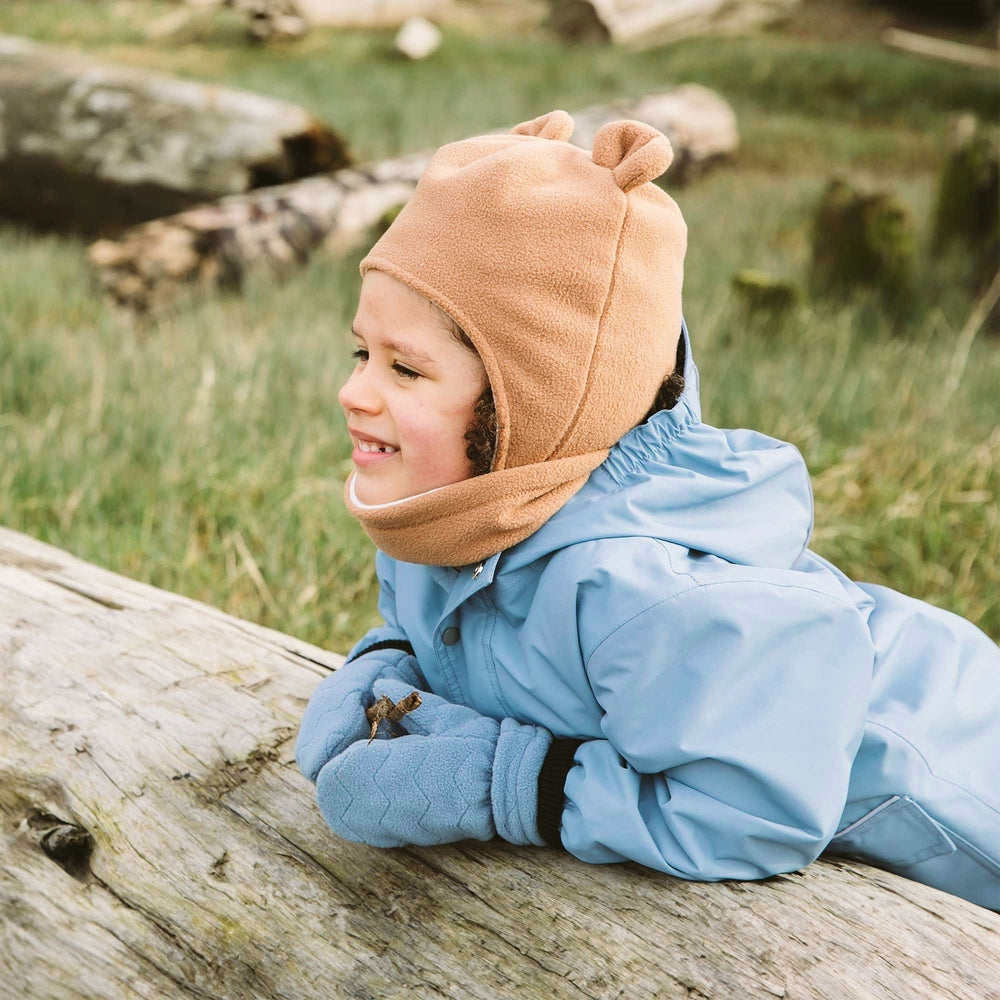 Jan & Jul Fleece Mittens Stormy Blue on a smiling child gripping a leaf while resting on a log outdoors.