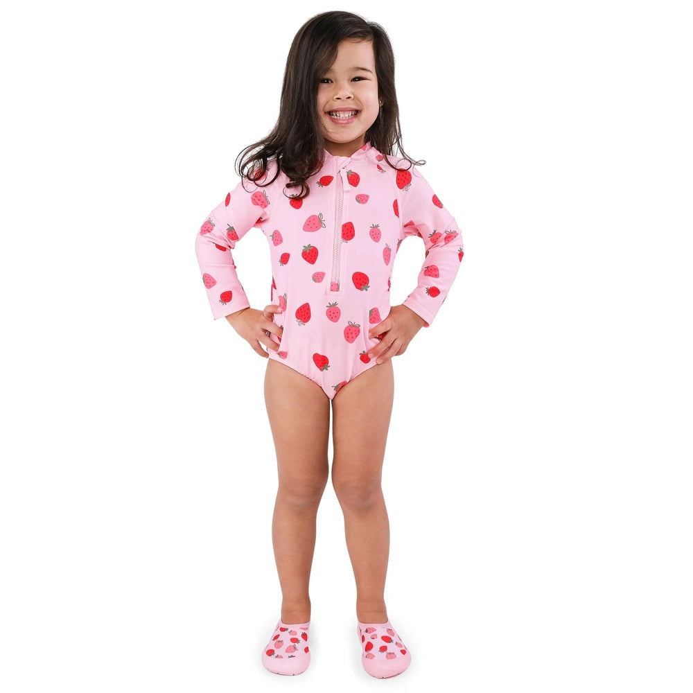 JAN & JUL girls one-piece UV swimsuit, pink strawberry print with front zipper, seen on a smiling girl posing with hands on hips.