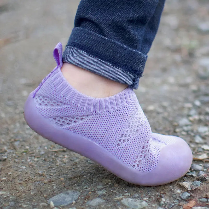 Jan & Jul Jelly Jumper Flow Shoes Lavender worn by a toddler walking on a gravel path, knit lavender upper.