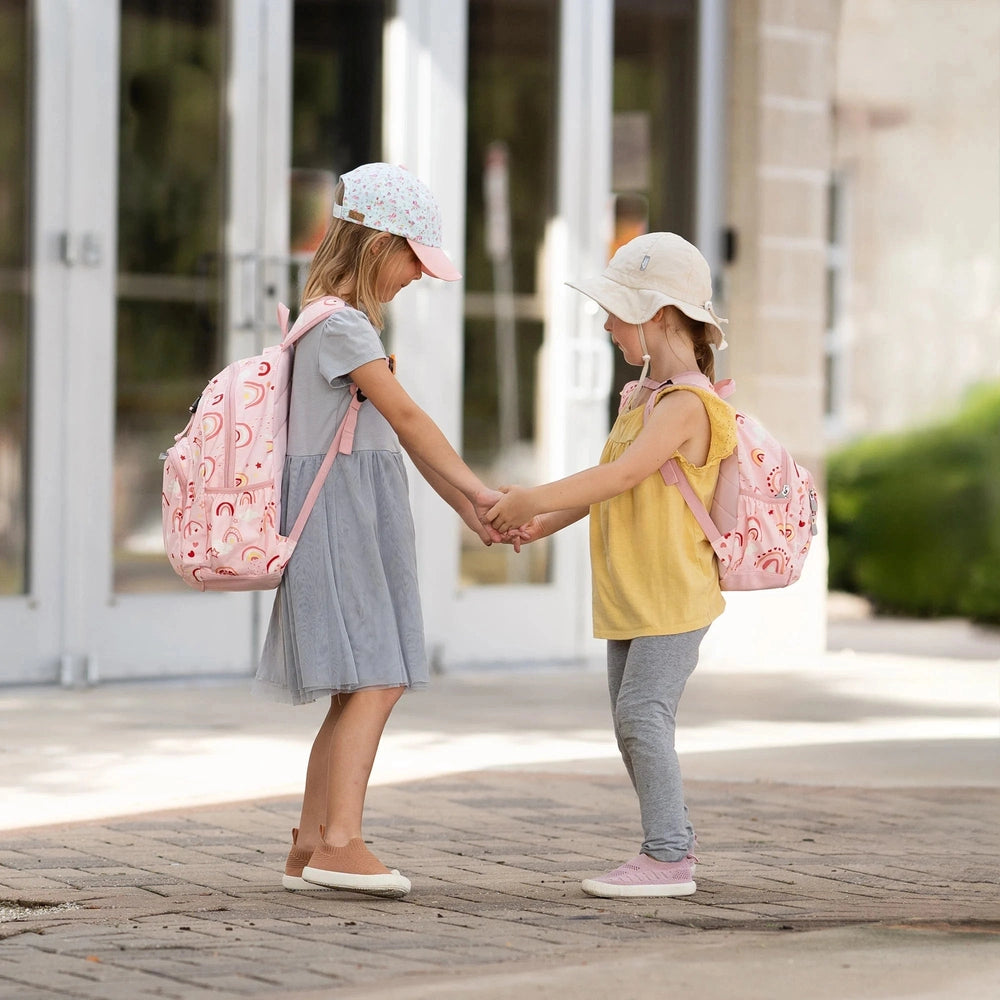 JAN & JUL Kids Backpacks in pink rainbow pattern worn by twin girls who hold hands, cap hats, showing cute backpacks.