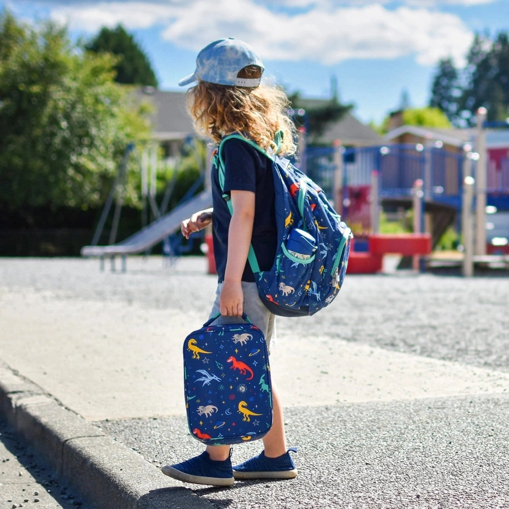 Jan & Jul Kids Backpack in blue dinosaur print with teal straps, worn by a child at the playground with a matching lunch bag.