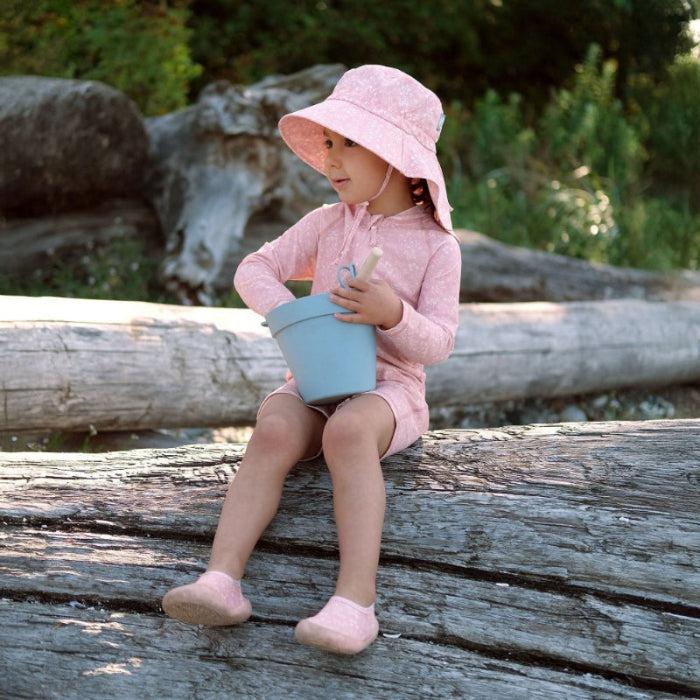 Jan & Jul Kids Cotton Adventure Hats Pink Prairie; toddler girl sits on a log in a pink wide-brim hat, holding a blue bucket.