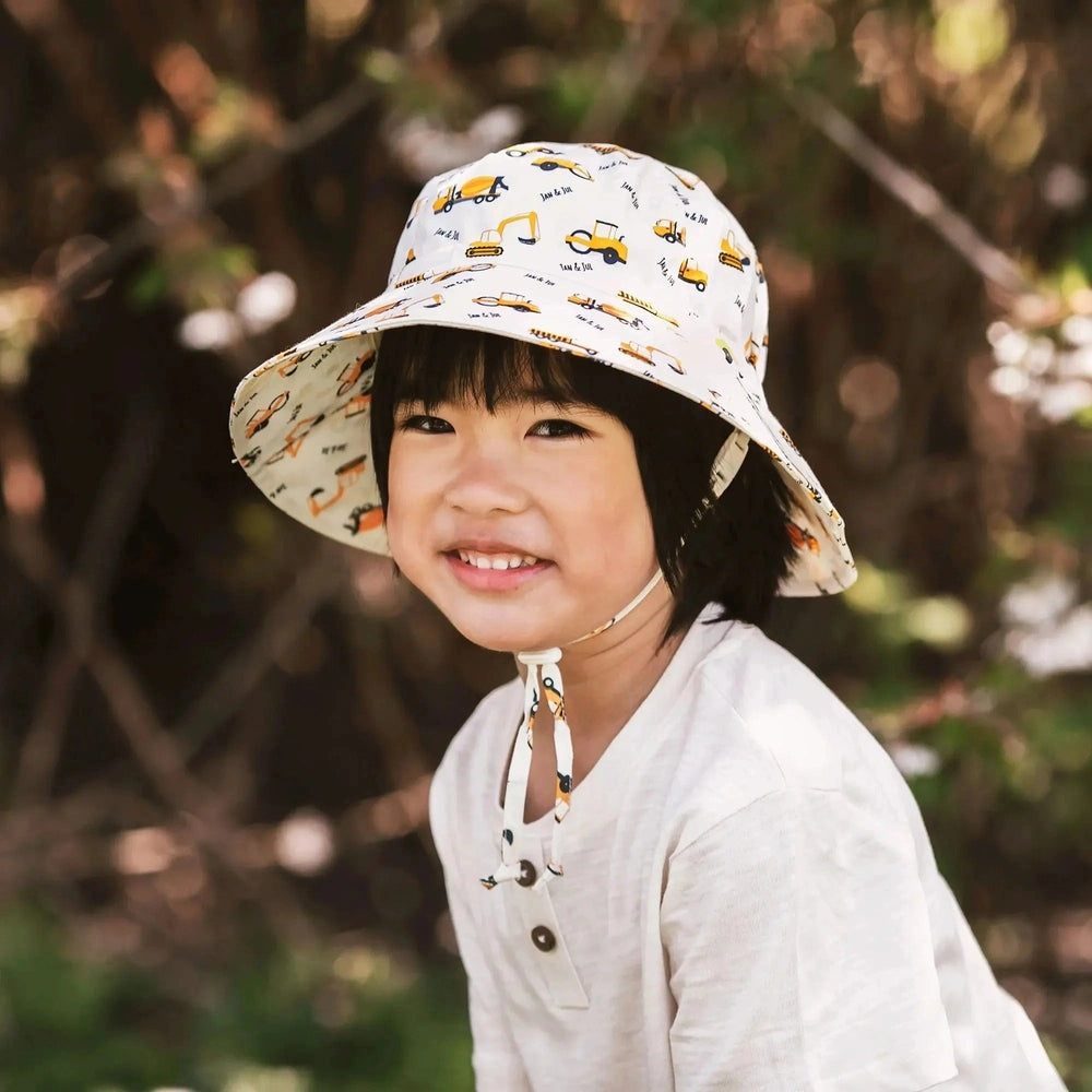 Jan & Jul Kids Cotton Bucket Hat Little Diggers, white with yellow construction vehicles, chin-strap under chin on smiling child