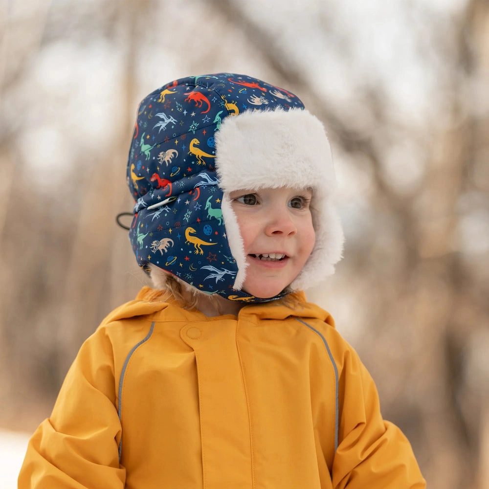 Jan & Jul Kids Insulated Winter Hats Space Dinos navy hat with white faux-fur trim worn by a smiling child outdoors in a mustard jacket.