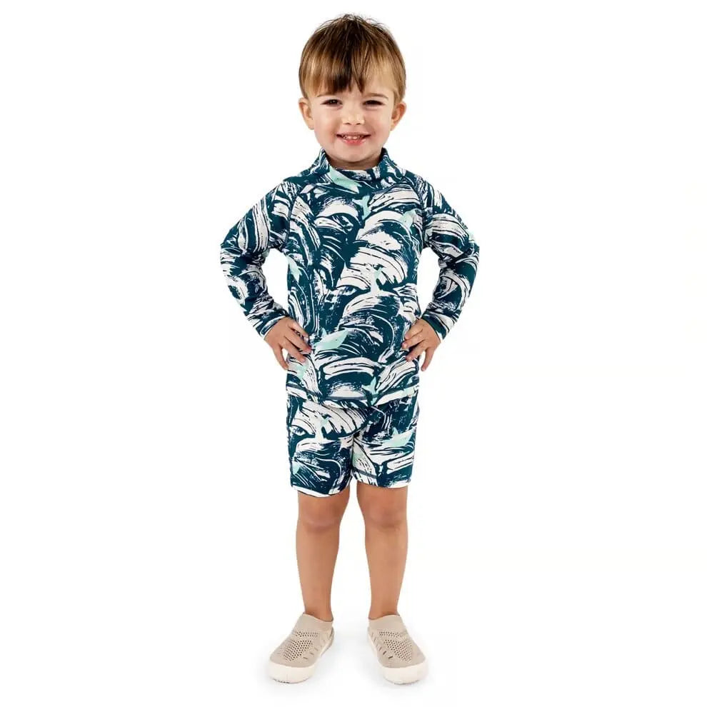 Jan & Jul Kids UV Rashguard Set Wave Rider teal palm print; boy stands smiling with hands on hips in matching long-sleeve top and shorts