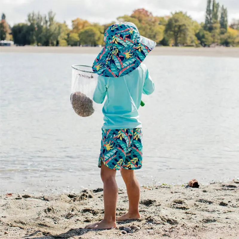 JAN & JUL Kids UV Rashguard Top Lagoon in turquoise, worn with tropical-print shorts by a sunny beach.