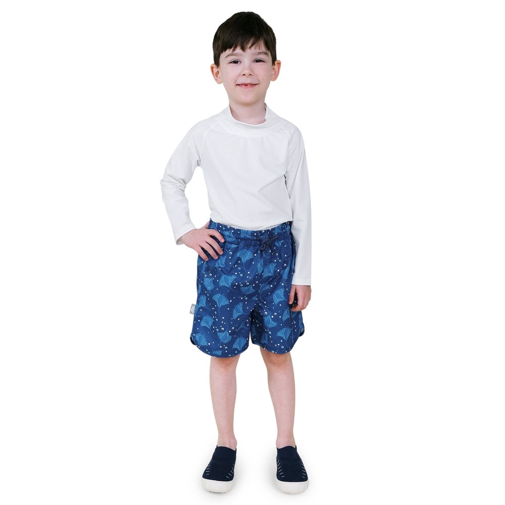 Jan & Jul Kids UV Swim Shorts Manta Ray in blue with manta ray pattern, worn by a smiling boy standing with hand on hip.