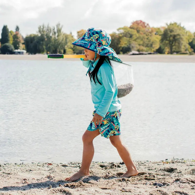 Jan & Jul Kids Water Repellent Adventure Hat Tropical worn by a child strolling a sandy beach in teal and tropical print.
