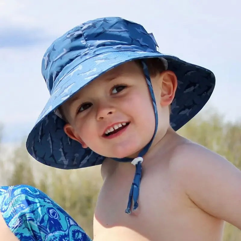 Jan & Jul Kids Water Repellent Bucket Hat Shark in blue with chin strap under toddler's chin on a sunny beach.
