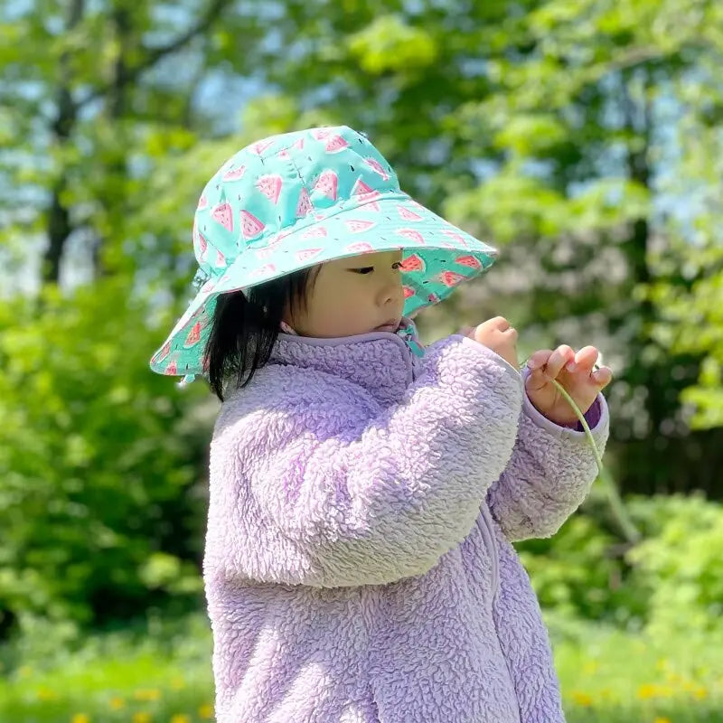 JAN & JUL Kids Water-Repellent Watermelon Bucket Hat in turquoise with watermelon print, toddler outdoors adjusting the strap