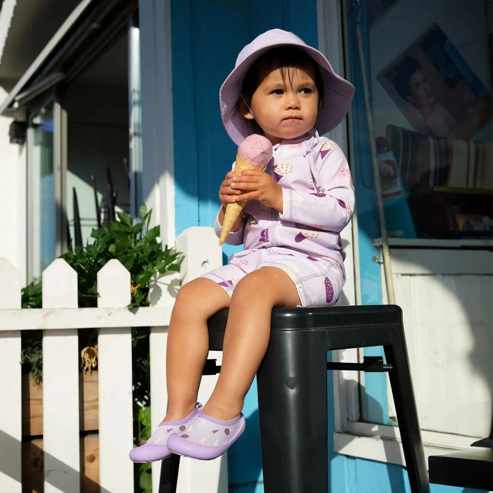 Jan & Jul Kids Water Shoes Lavender Ice Cream worn by a toddler sitting on a stool and holding an ice cream cone.