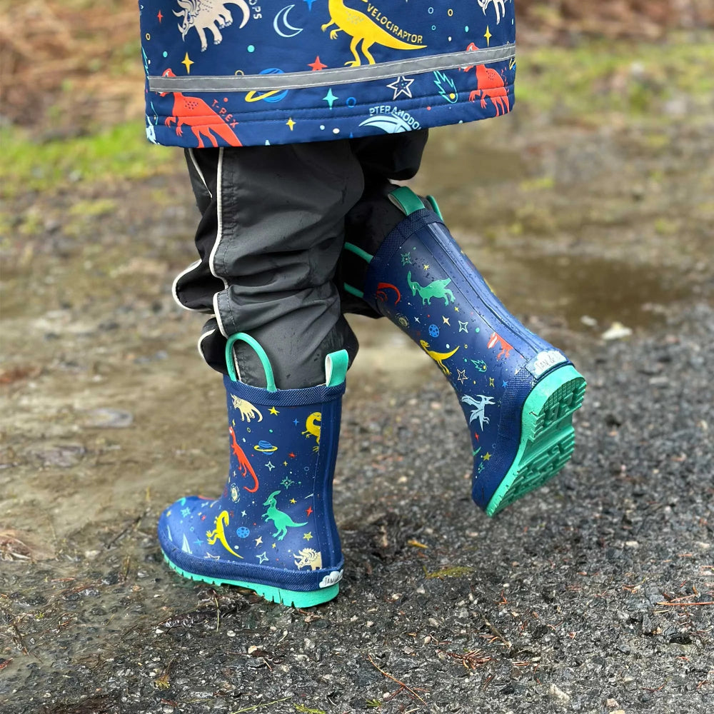 Jan & Jul Puddle Dry Rain Boots Space Dinos for toddlers, blue with green soles, a child splashing through a wet path