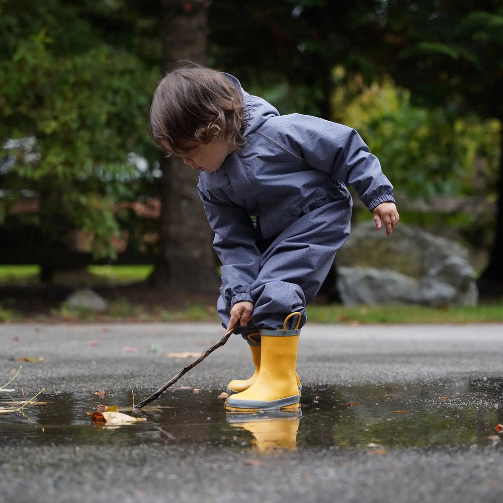 Toddler in a blue rain suit wearing Jan & Jul Puddle Dry Rain Boots With Handle Yellow, using a stick to poke a puddle.