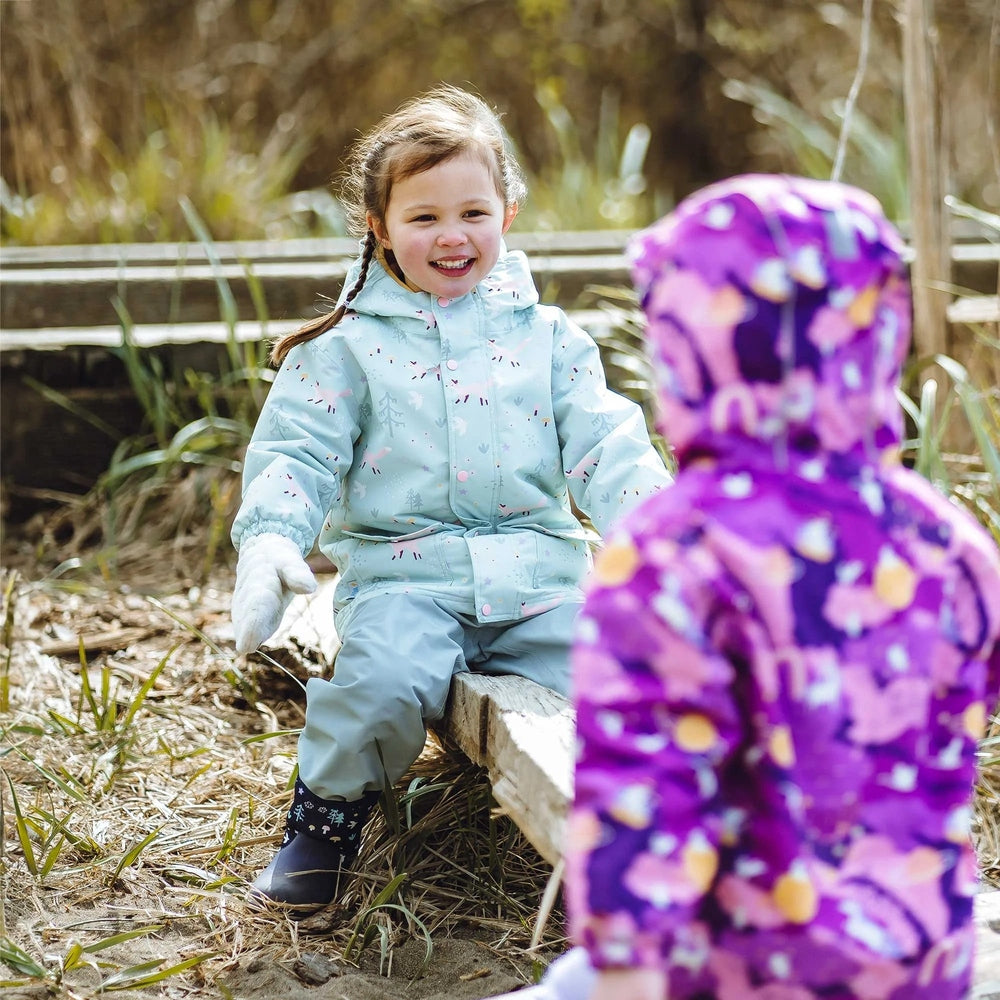 JAN & JUL Waterproof Cozy Dry Rain Jacket Purple Dream worn by a smiling girl playing outdoors on wooden steps.