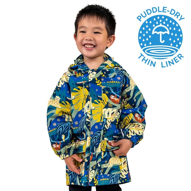 JAN & JUL Waterproof Puddle Dry Rain Jacket Jungle Nights on a smiling child in blue jungle print, hood up, hands in pockets