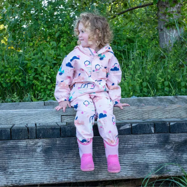 Toddler in JAN & JUL Waterproof Puddle Dry Rain Pants Dreamscape pink rain suit sits on a wooden bench outdoors.