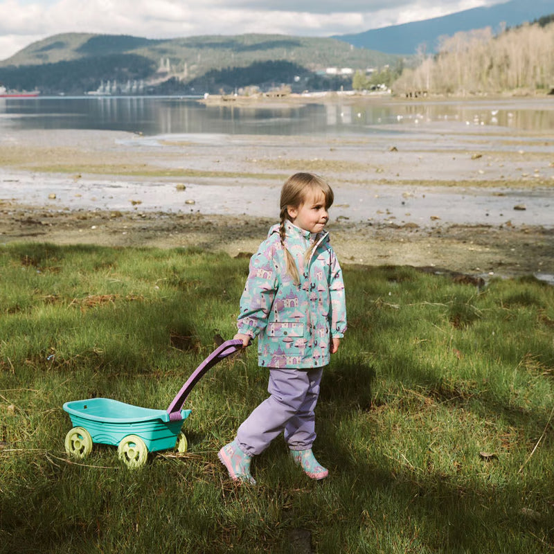 JAN & JUL Lavender waterproof puddle-dry rain pants worn by a toddler pulling a turquoise wagon along a grassy shore.