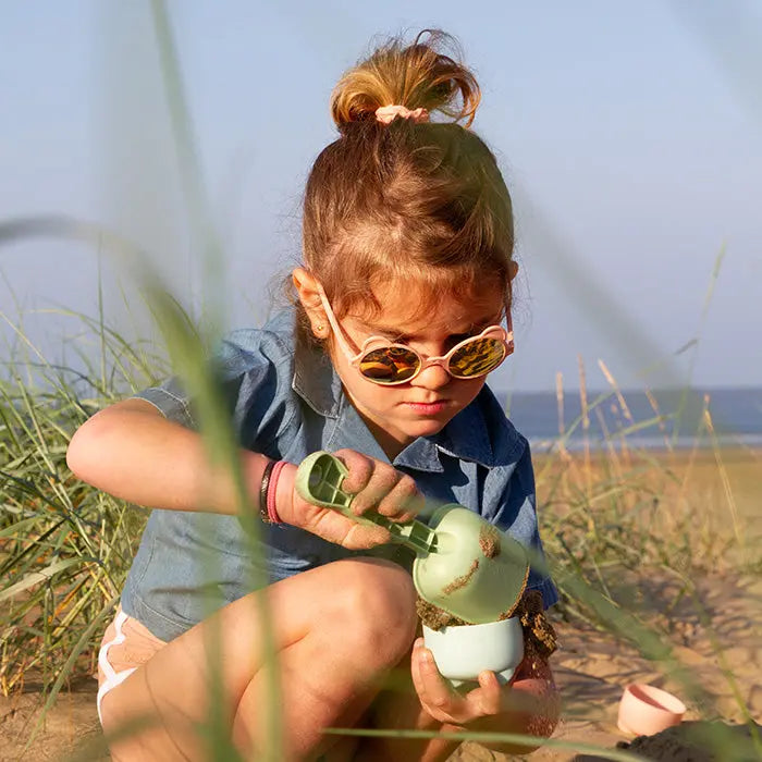 Ki Et La Sunglasses Ourson Peach Pink on a girl at the beach, showing round peach-pink frames as she dumps sand into a cup