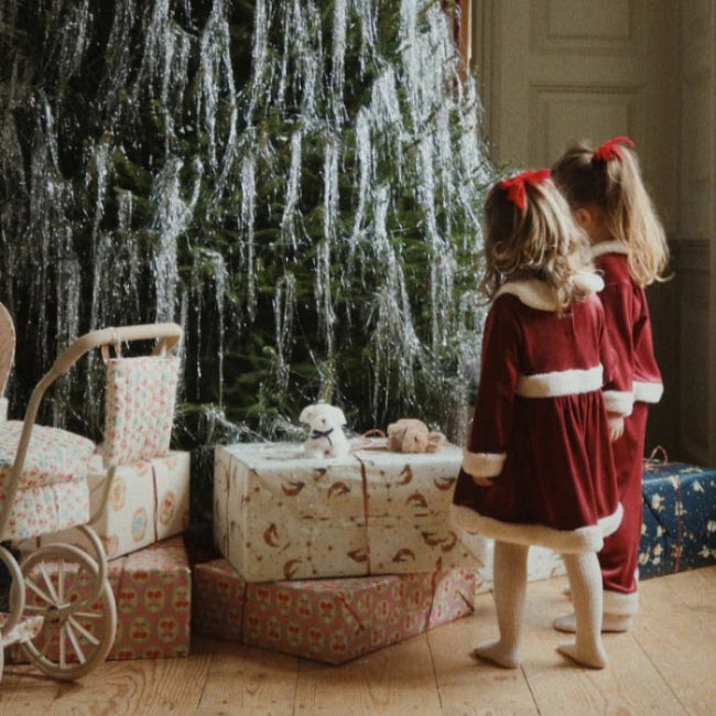 Konges Slojd Christmas Dress Red Dahlia for toddlers, two girls in red velvet with white fur trim stand by wrapped gifts.