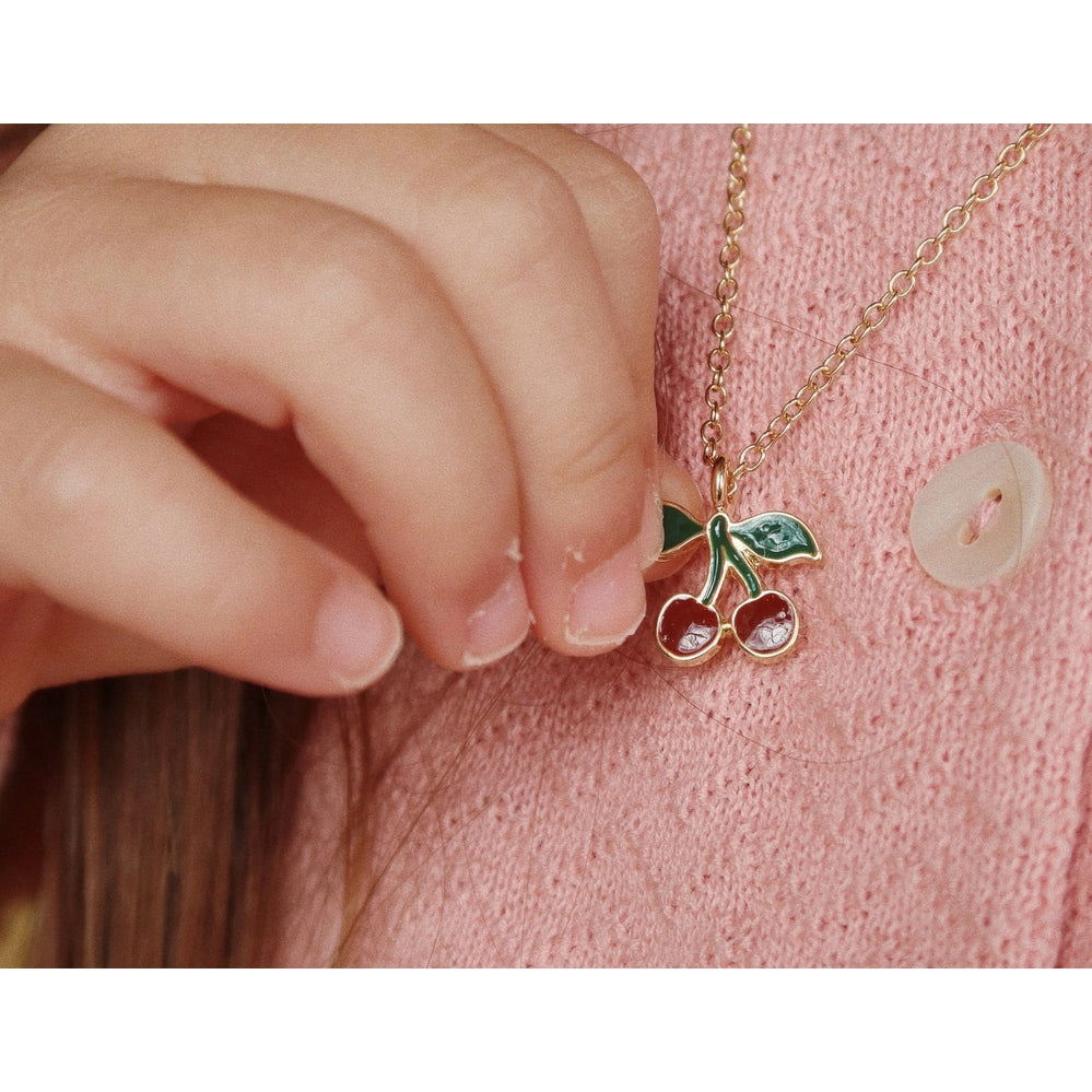 Konges Slojd Jewelry Set cherry pendant necklace held by a child's fingers against a pink knit cardigan.
