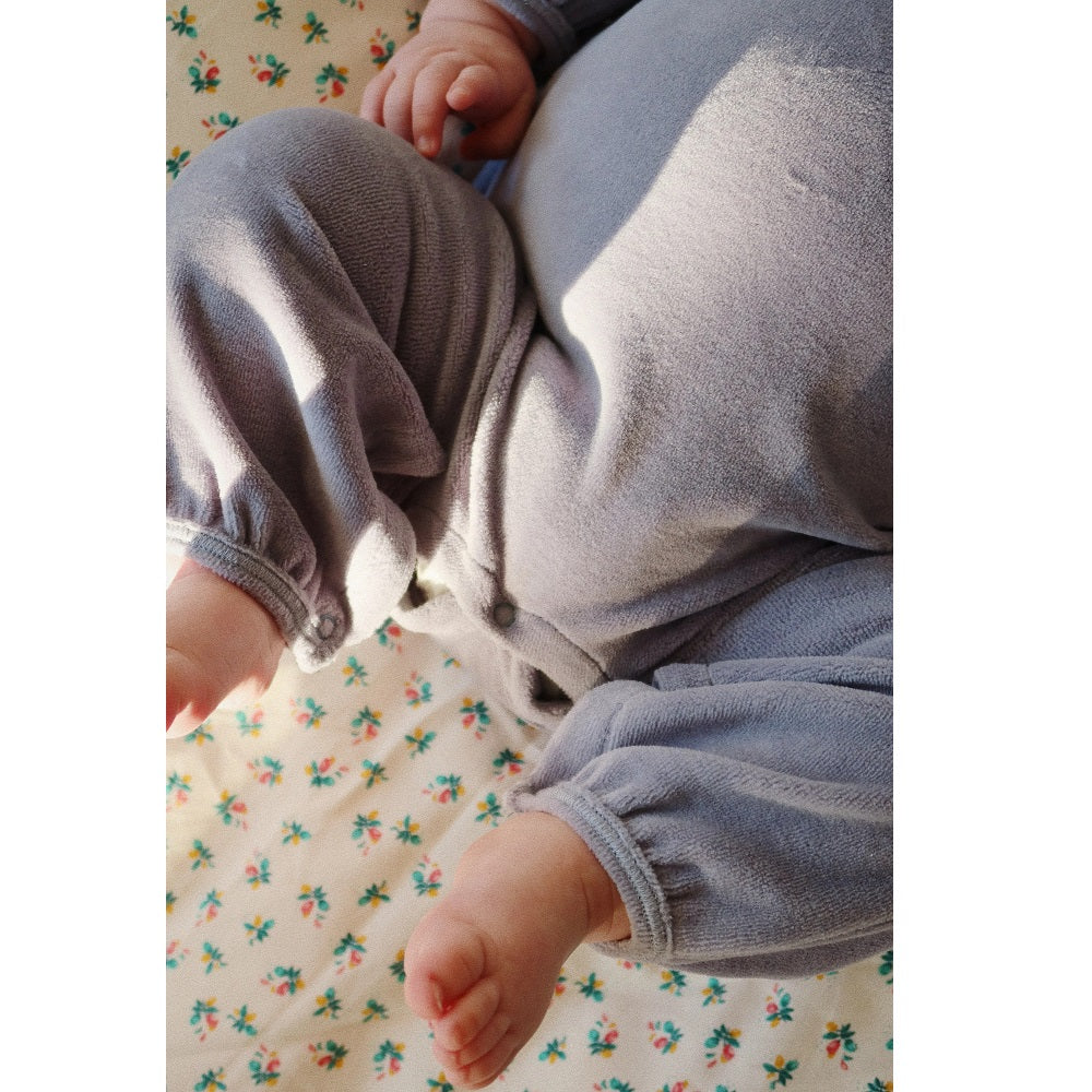Konges Slojd Miffa Onesie Trade Winds gray-blue fleece; baby's feet and hands rest on a floral blanket.