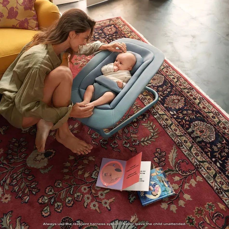 Lifestyle image of the Cybex Lemo 2 Bouncer in blue, where a mother leans in as the baby sits securely harnessed.