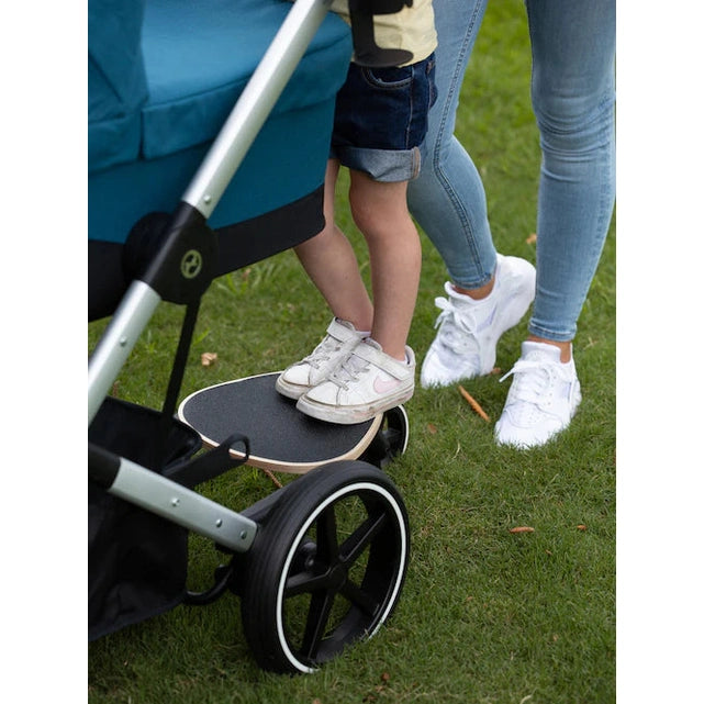 Lifestyle image of the Cybex Platinum Accessories Kid Board with a child standing on the black board attached beside a stroller rear wheel on grass.
