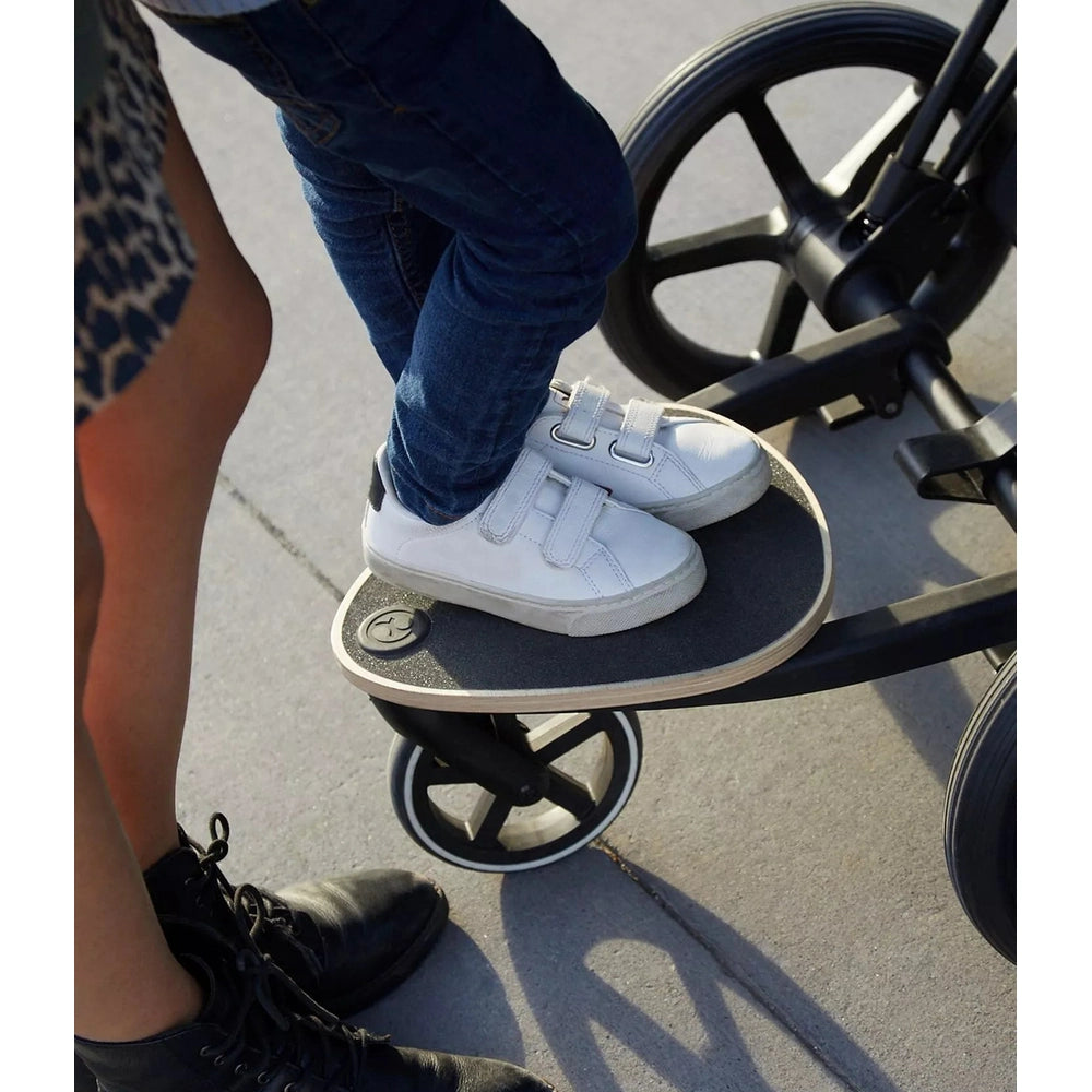 Lifestyle image of the Cybex Platinum Accessories Kid Board with a child standing on the black ride-on board beside a stroller wheel on sidewalk.