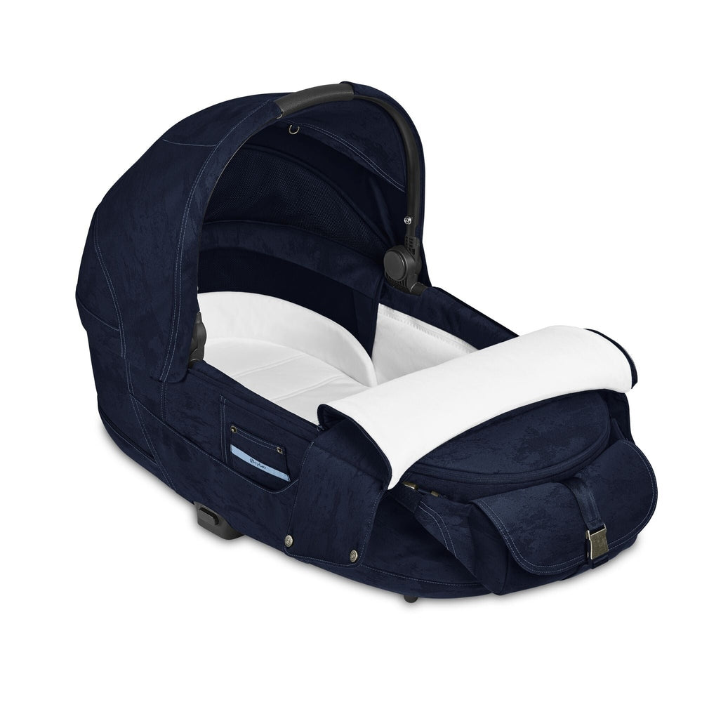 Lifestyle image of the Cybex Platinum Priam Carry Cot Limited Edition - Rebellious Luxury Denim Blue navy blue denim carry cot for stroller with white padded interior and curved canopy.