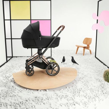 Lifestyle image of the Cybex Platinum Priam Lux Carry Cot stroller with black bassinet on a rose-gold frame, displayed on a wooden platform.