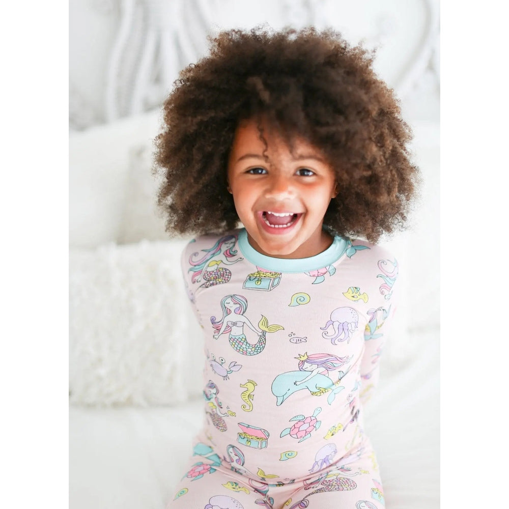 Little Sleepies Bamboo Pajama Set Mermaid in pastel pink; smiling child sits on a bed wearing mermaid-print pajamas.