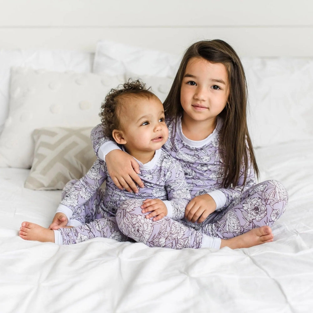 Little Sleepies Bamboo Pajama Set Peonies in lavender florals; sister hugs baby on a white bed.