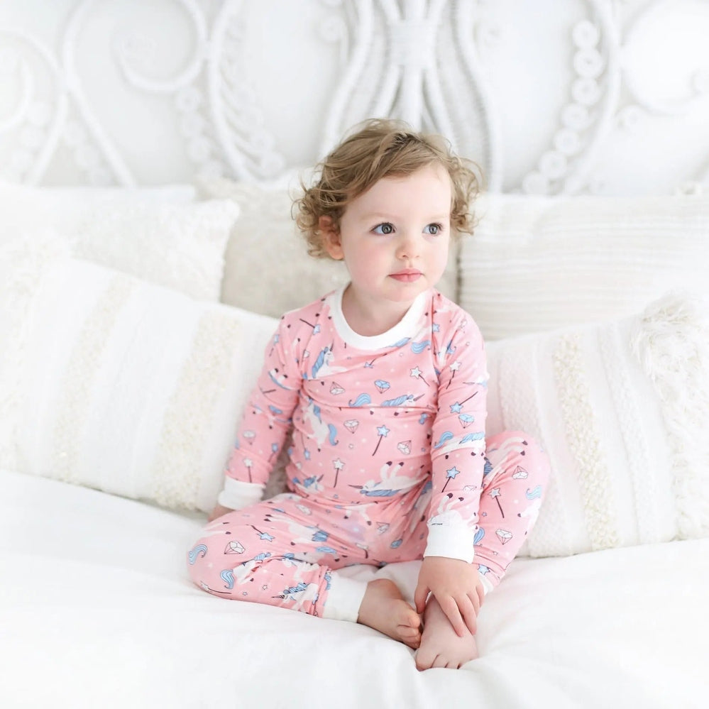 Little Sleepies Bamboo Pajama Set Pink Unicorn; toddler girl sits on a white bed in a cozy, bright all-white bedroom.