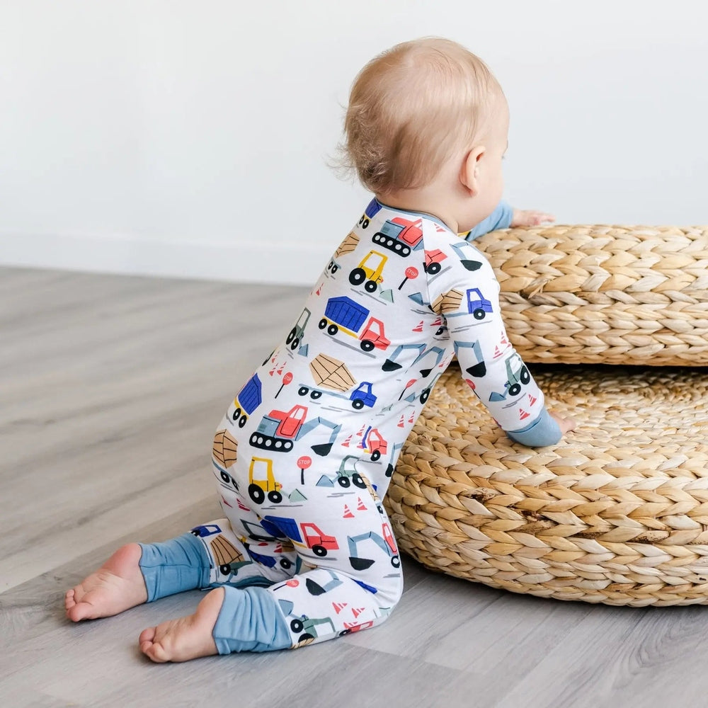 Littlesleepies Construction Bamboo Viscose Zippy: white onesie with colorful trucks, baby crawls on floor beside woven pouf.