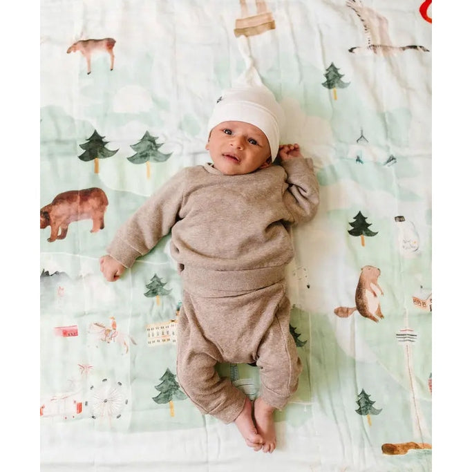 Loulou Lollipop Muslin Quilt Blanket; baby in beige lays on pastel forest scene blanket, showcasing soft muslin swaddle.