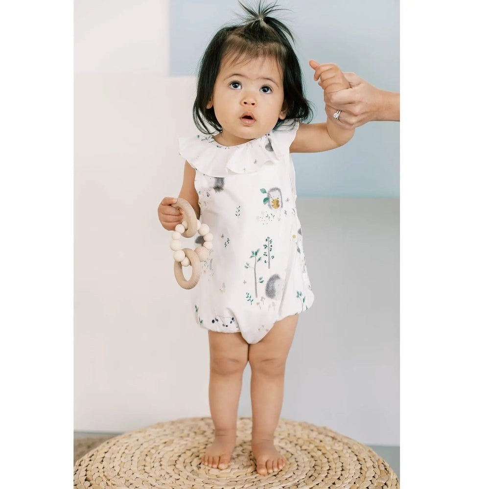 Loulou Lollipop Ruffle Bubble Suit Hedgehogs in white; baby stands on a woven mat, gripping a wooden teether as an adult guides her.