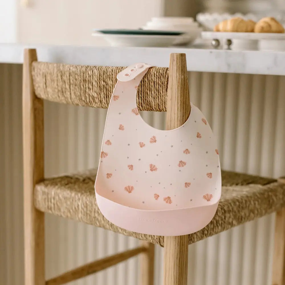 Loulou Lollipop Silicone Bib pale pink with shell print, adjustable strap snapped onto a wooden chair to catch spills