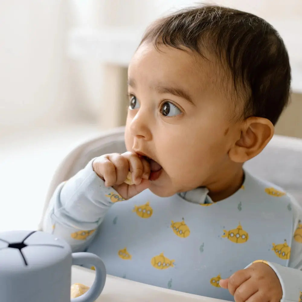 Loulou Lollipop Silicone Bib, light-blue with yellow submarine print, catching crumbs as baby eats.