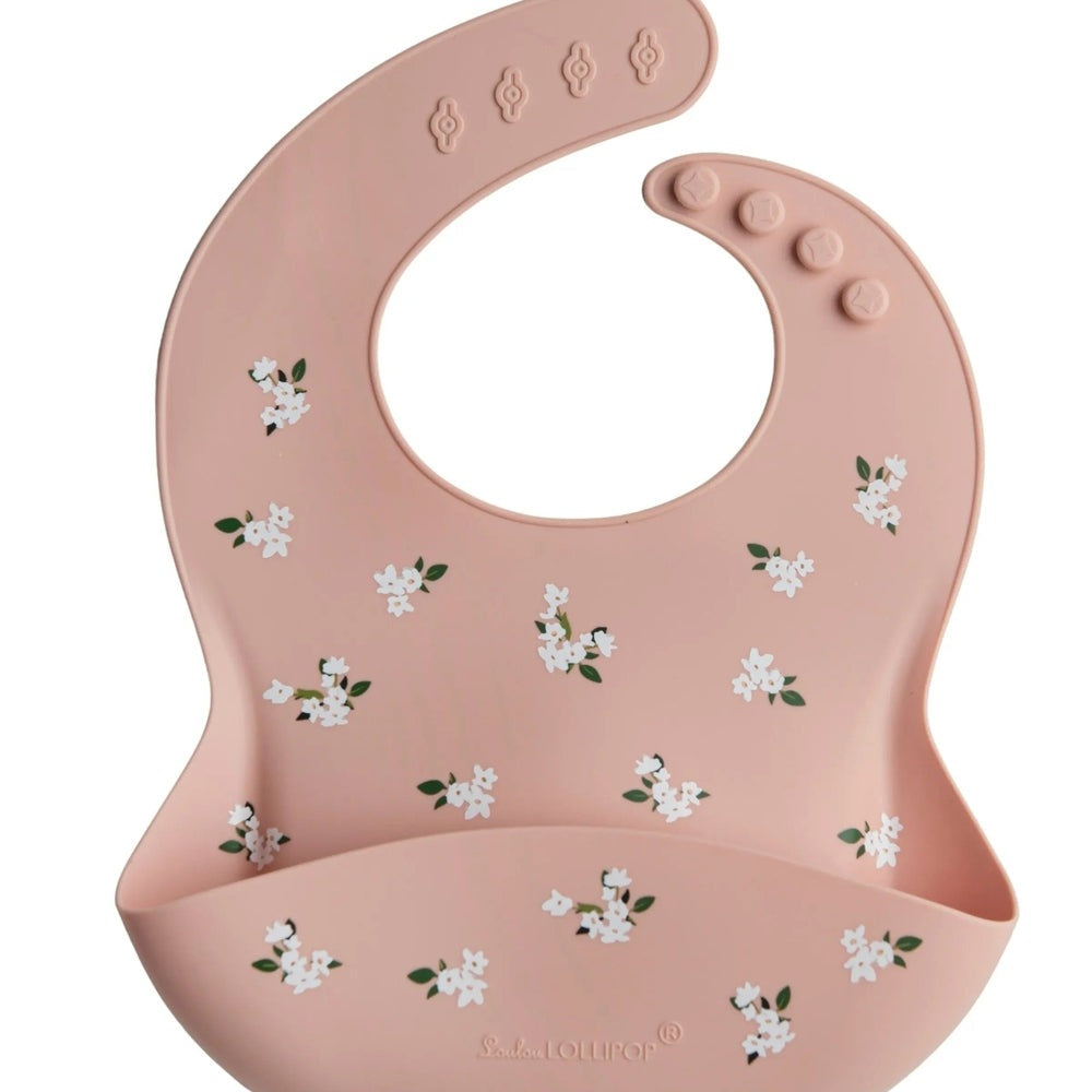 Loulou Lollipop silicone bib in pink with white flowers; large front pocket catches crumbs during feeds.