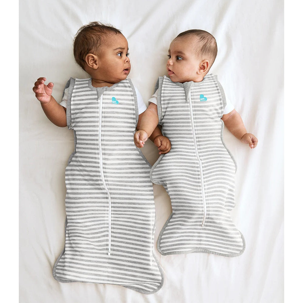 LOVE TO DREAM Swaddle Up Transition Bag Original Cotton 1.0 Tog Grey; twins lie on a white sheet in striped sleep sacks with front zippers.